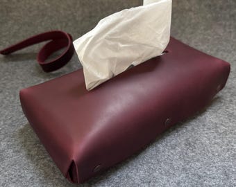 May include: A burgundy leather tissue box with a rectangular shape and a strap. White tissues are pulled from the top opening. The box has metal rivets on the side.