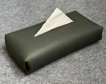 May include: A rectangular tissue box cover in a dark green hue. A single, folded white tissue protrudes from the top opening. The box has a smooth, matte finish, suggesting a modern design. The tissue box is placed on a gray surface.
