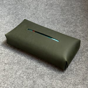 May include: An olive green rectangular tissue box cover with a rectangular opening on the top. The tissue box cover is made of a smooth material and sits on a gray surface.