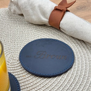 Puede incluir: Un posavasos de cuero azul oscuro con un borde cosido azul. El posavasos está personalizado con el nombre "Brown" en una fuente cursiva.