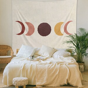 May include: A beige tapestry with a minimalist design featuring three crescent moons in shades of red, orange, and yellow, and a large circle in maroon. The tapestry is hanging on a wall above a bed with white bedding and pillows.
