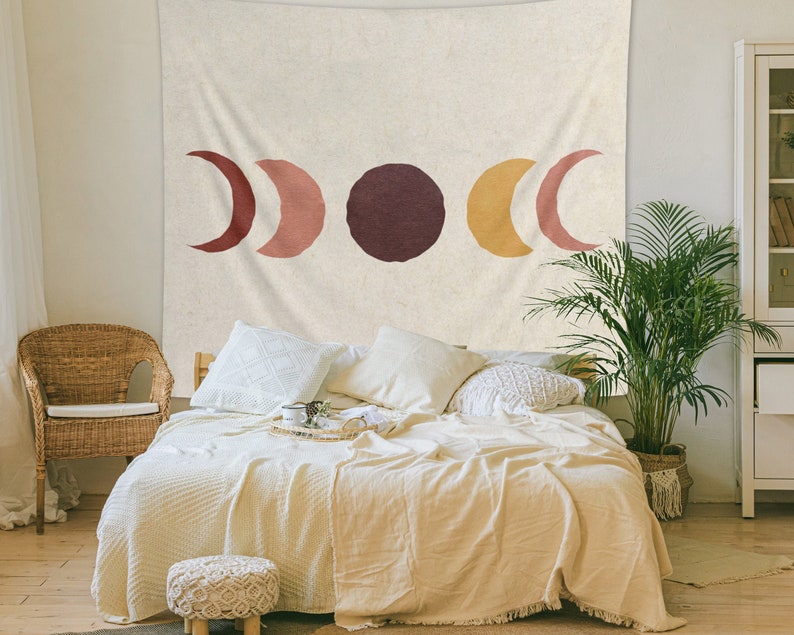 May include: A beige tapestry with a minimalist design featuring three crescent moons in shades of red, orange, and yellow, and a large circle in maroon. The tapestry is hanging on a wall above a bed with white bedding and pillows.