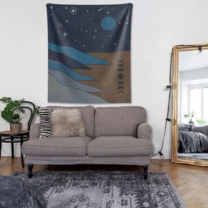 May include: A gray couch with two pillows, a throw blanket, and a tapestry hanging on the wall behind it. The tapestry features a blue and brown abstract design with stars, a crescent moon, and a full moon.