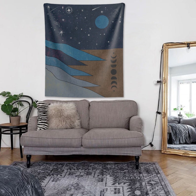 May include: A gray couch with two pillows, a throw blanket, and a tapestry hanging on the wall behind it. The tapestry features a blue and brown abstract design with stars, a crescent moon, and a full moon.