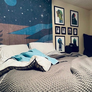 May include: A gray knitted blanket on a bed with white pillows. The bed is in front of a wall with framed artwork featuring black and white illustrations of people. A tapestry with a blue and brown mountain scene hangs on the wall behind the bed.