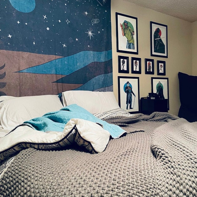 May include: A gray knitted blanket on a bed with white pillows. The bed is in front of a wall with framed artwork featuring black and white illustrations of people. A tapestry with a blue and brown mountain scene hangs on the wall behind the bed.
