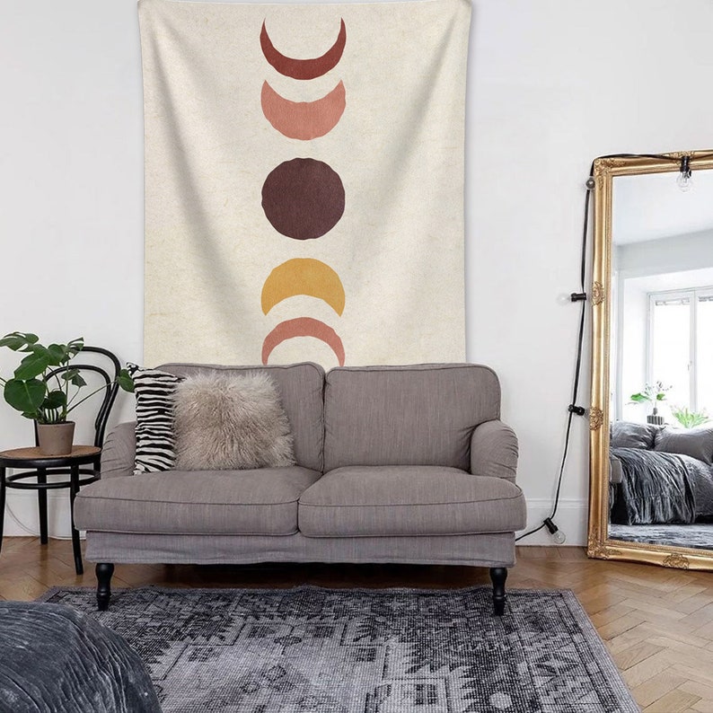 May include: A beige tapestry with a vertical design of five abstract moons in shades of red, pink, brown, and yellow. The tapestry is hanging on a wall above a gray couch with two pillows.