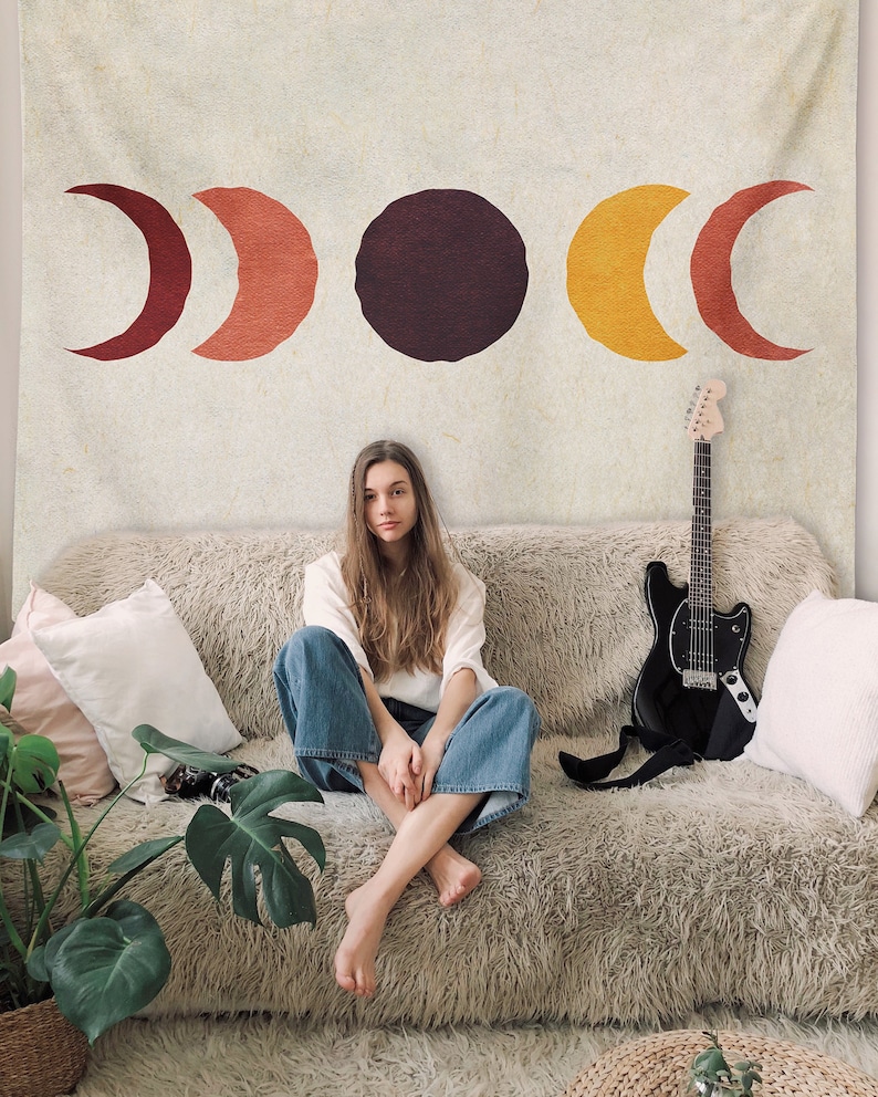 May include: A tapestry with a design of three moons in different phases, with a red, orange, and yellow color scheme. The tapestry is hanging on a wall behind a couch with a person sitting on it.