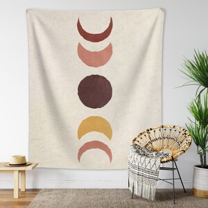May include: A tapestry with a beige background and a vertical arrangement of five abstract shapes. The shapes are in shades of brown, orange, and pink, resembling moons and circles.