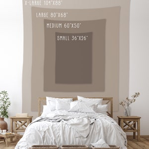 May include: A white bed with a white blanket and pillows, a brown wooden headboard, and two wicker baskets at the foot of the bed. The bed is in front of a wall with a beige tapestry hanging on it. The tapestry has text that reads "X-LARGE 101"x88", "LARGE 80"x68", "MEDIUM 60"x50", and "SMALL 36"x26".