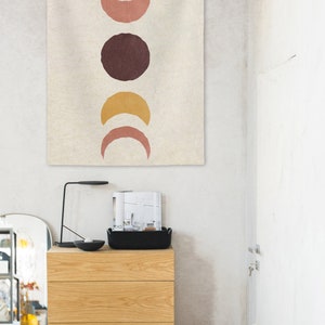 May include: A beige tapestry with a vertical row of five abstract moon shapes in shades of brown, pink, and yellow. The tapestry is hanging on a white wall.