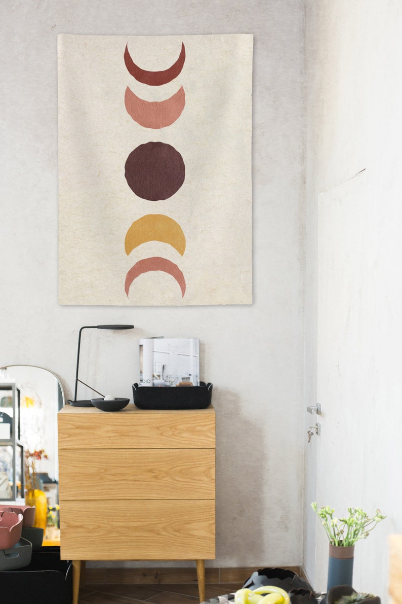 May include: A beige tapestry with a vertical row of five abstract moon shapes in shades of brown, pink, and yellow. The tapestry is hanging on a white wall.
