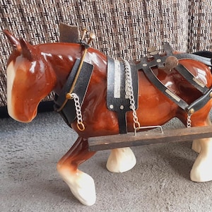 May include: A ceramic brown horse figurine with white markings on its face and legs, wearing a black harness with silver chains. The horse is attached to a wooden cart carrying four dark barrels with gold-coloured bands and a large black wheel.