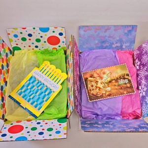 Pu&ograve; includere: Due scatole regalo aperte con motivi colorati a pois e fiocchi di neve. Una scatola contiene un biglietto "Happy Birthday!" e carta velina gialla e verde. L'altra scatola ha un biglietto "Sweet tidings" e carta velina viola e rosa.