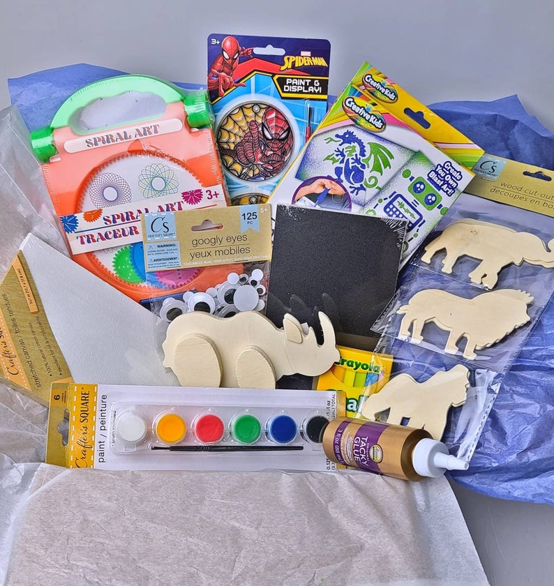 Pu&ograve; includere: Una collezione di materiali per l'artigianato tra cui un kit di arte a spirale, un set di vernici, occhi finti, sagome di animali in legno e un kit di pittura e esposizione di Spider-Man. Gli articoli sono disposti in una scatola con carta velina blu.