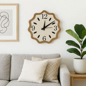 May include: A decorative wall clock with a light beige face and a scalloped wooden frame. Black numbers and markers indicate the time. The clock is mounted on a white wall above a grey sofa with pillows. A framed abstract art piece is to the left.