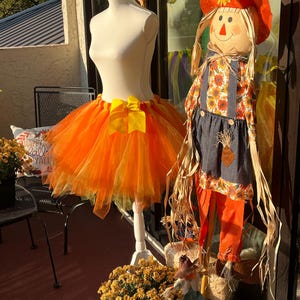 May include: An orange and yellow tulle tutu skirt with a yellow bow is displayed on a mannequin. A scarecrow decoration with a sunflower hat and overalls stands nearby. Fall-themed decor.