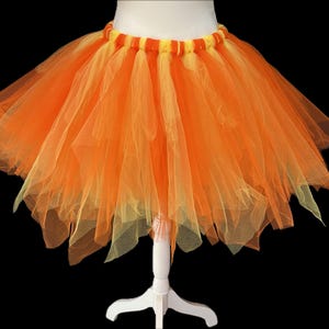 Peut inclure: Un tutu orange et jaune vif, présenté sur un mannequin blanc. La jupe est faite de couches de tulle, créant un aspect ample et duveteux. La ceinture est un mélange d'orange et de jaune. Parfait pour les costumes ou la danse.