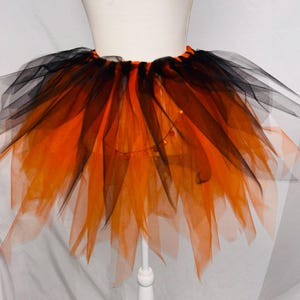 May include: A layered tulle tutu skirt featuring alternating strips of vibrant orange and black fabric. The skirt has a gathered elastic waistband and a voluminous, flared silhouette with uneven, pointed hem edges throughout the design.