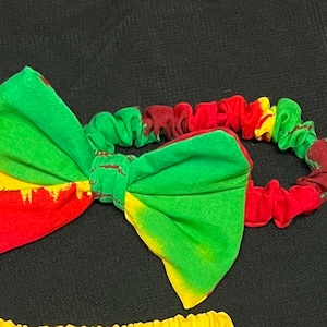 Puede incluir: Una diadema de tela verde, amarilla y roja con un lazo. El lazo está hecho de una tela vibrante con un patrón audaz. La diadema es de estilo scrunchie, con la misma combinación de colores que el lazo.