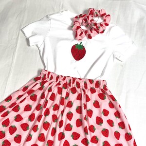 Op de afbeelding: Een wit t-shirt met een grote geborduurde aardbei, een roze rok met aardbeienpatroon en twee roze scrunchies met aardbeienprints. De rok en scrunchies hebben een roze achtergrond met rode aardbeien.