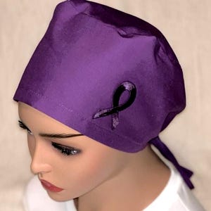 May include: A solid purple scrub cap featuring a dark, textured awareness ribbon appliqué on the side. The cap is constructed with a fitted crown and includes fabric ties at the back for adjustment, providing a secure and comfortable fit for wearers.