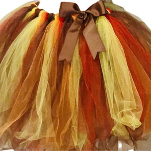 Puede incluir: Una falda tutú en capas en colores otoñales, que incluye tonos de marrón, naranja y amarillo. La falda está hecha de tul y tiene un lazo de satén marrón en la cintura. La falda está diseñada para niños.