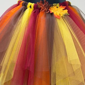 Peut inclure: Jupe tutu multicolore aux couleurs automnales. La jupe présente des couches de tulle orange, jaune, marron et bordeaux. Une feuille décorative et une tige sont attachées à la ceinture. Idéale pour les costumes ou les événements à thème.
