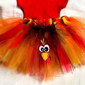 Puede incluir: Un body rojo de manga corta unido a una falda de tutú de tul de varias capas en tonos rojo, naranja, amarillo y marrón. La falda presenta una aplicación bordada de cara de pavo con ojos blancos, un pico amarillo y una carúncula roja.