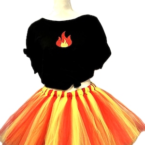 Op de afbeelding: Een zwart shirt met korte mouwen en een centraal rood en geel vlammotief op de borst, gecombineerd met een gelaagde tule tutu-rok met afwisselende verticale strepen van felrode, oranje en gele stof over het gehele oppervlak.