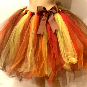 May include: A colorful tutu skirt in shades of brown, yellow, orange, and red. The skirt is made of layered tulle and features a brown satin bow at the waist. This festive skirt is suitable for costumes or special occasions.