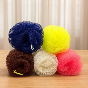 May include: A collection of six colorful mesh bath sponges in shades of blue, yellow, brown, white, and pink. The sponges are arranged on a wooden surface, with a neutral background.