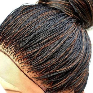 May include: A black and brown braided wig with a lace front. The wig is styled in a high bun.