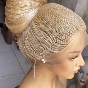 May include: A blonde braided wig styled in a high bun. The wig is on a mannequin head, showcasing the intricate braiding and the natural-looking hairline. The wig is a light, golden color, and the braids are tightly woven.