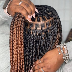 May include: A woman with brown and black box braids. The braids are styled in a half-up, half-down style. The woman is wearing a silver bracelet on her wrist.