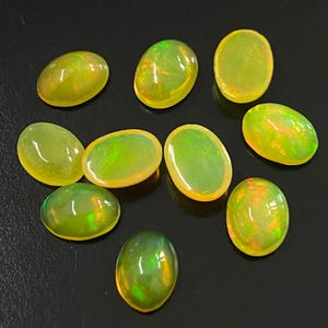 Op de afbeelding: Een verzameling van tien ovale opalen. De opalen hebben een doorschijnend uiterlijk met een mix van gele, groene en oranje tinten. De edelstenen zijn gerangschikt op een donkere ondergrond, wat hun iriserende kwaliteiten benadrukt.
