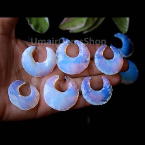 May include: A selection of opalescent, crescent-shaped gemstones. The stones display a milky white base with iridescent blue and purple tones. The gemstones are presented on a dark background, highlighting their unique shape and colour variations.