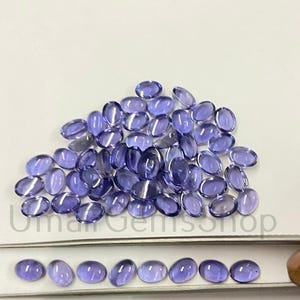 Puede incluir: Una colección de gemas ovaladas, translúcidas, de color azul violeta. Las gemas están dispersas sobre una superficie blanca, con algunas dispuestas en fila. El texto "Umar Gemshop" es visible.