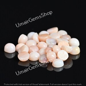 May include: A collection of polished, oval-shaped gemstones in shades of peach and cream. The stones are smooth and reflective, with a glossy finish. The image is set against a dark background, highlighting the gems.
