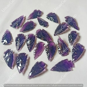 May include: A collection of 20 purple amethyst arrowhead points. The arrowheads are various sizes and shapes, with some having a more pointed tip than others. The arrowheads are all a deep purple colour with some having a lighter purple hue.
