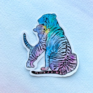 Puede incluir: Una ilustración de acuarela de dos tigres, uno más grande que el otro, abrazándose. Los tigres están pintados en tonos de azul, morado y rosa.