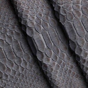 May include: Close-up of a textured, gray snakeskin pattern. The scales are oval-shaped and overlap, creating a detailed and realistic appearance. The folds in the material add depth and dimension to the image. This material is suitable for fashion and accessories.