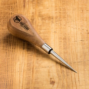 Puede incluir: Un punzón de cuero de madera con una punta de metal y un mango de madera marrón. El mango tiene un logotipo que dice "BANTAMA" con un gráfico de cabeza de toro.