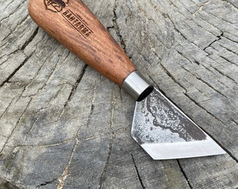 May include: A small, hand-forged knife with a wooden handle. The handle is engraved with the words "HAND MADE" and "BAKER TREE" along with a logo. The knife is resting on a wooden surface.