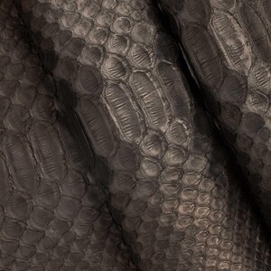 May include: Close-up of a dark gray, textured material with a scale-like pattern. The surface has a subtle sheen, with variations in tone creating depth and dimension. The material appears to be leather or a similar fabric.