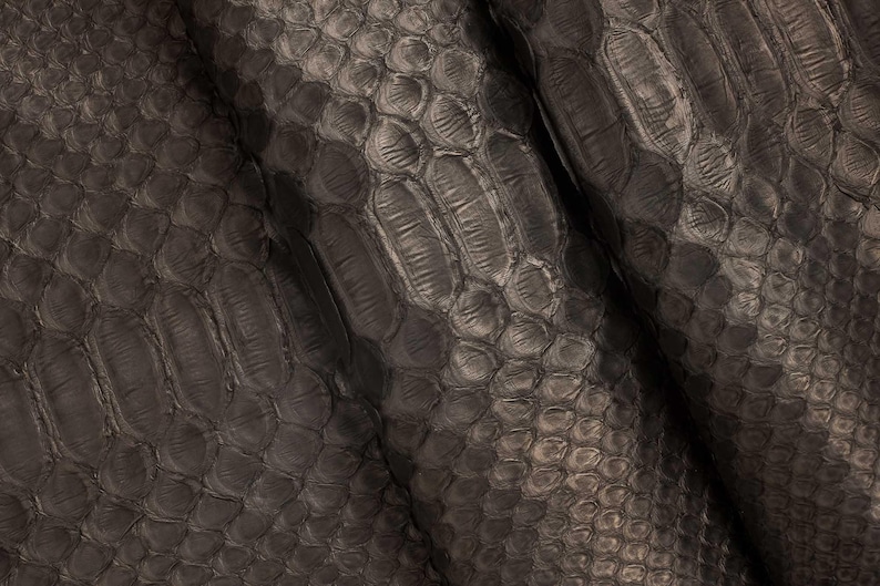 May include: Close-up of a dark gray, textured material with a scale-like pattern. The surface has a subtle sheen, with variations in tone creating depth and dimension. The material appears to be leather or a similar fabric.