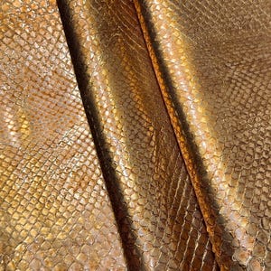 May include: Close-up of a golden, textured material with a snakeskin pattern. The fabric has a metallic sheen, with variations in color from light gold to darker bronze. The material is folded, creating shadows and highlighting the texture.