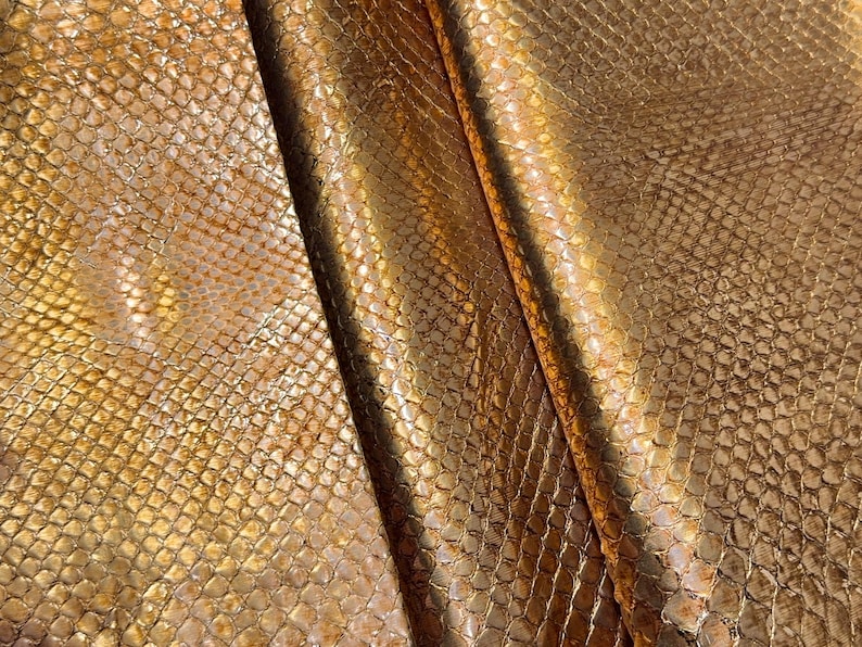 May include: Close-up of a golden, textured material with a snakeskin pattern. The fabric has a metallic sheen, with variations in color from light gold to darker bronze. The material is folded, creating shadows and highlighting the texture.