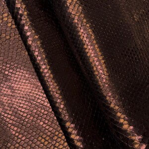 May include: Close-up of a textured, dark brown material with a snakeskin pattern. The material has a subtle sheen and a gradient of colors, including shades of brown, pink, and gold. The scales are small and closely packed, creating a detailed surface.