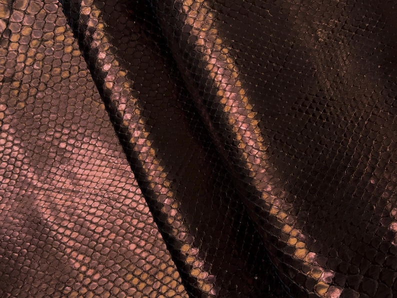 May include: Close-up of a textured, dark brown material with a snakeskin pattern. The material has a subtle sheen and a gradient of colors, including shades of brown, pink, and gold. The scales are small and closely packed, creating a detailed surface.